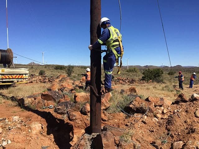 Harnessed linesman in hi-viz and hard hat climbing a steel pole on a wind farm collector line, ground crew and drum truck behind