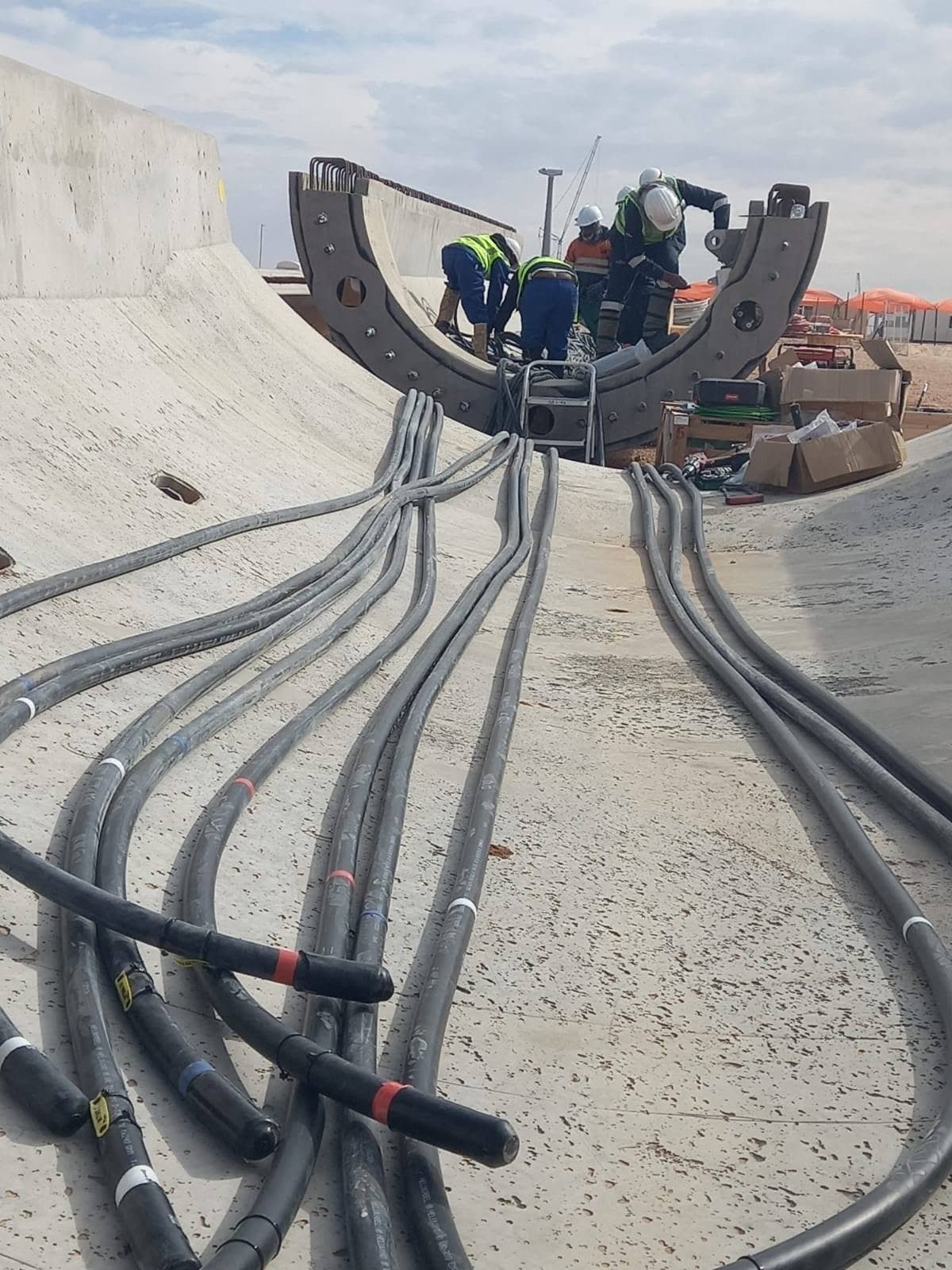 Fan of grey power cables sweeping across a concrete apron toward a curved wind turbine tower section where a crew is terminating them