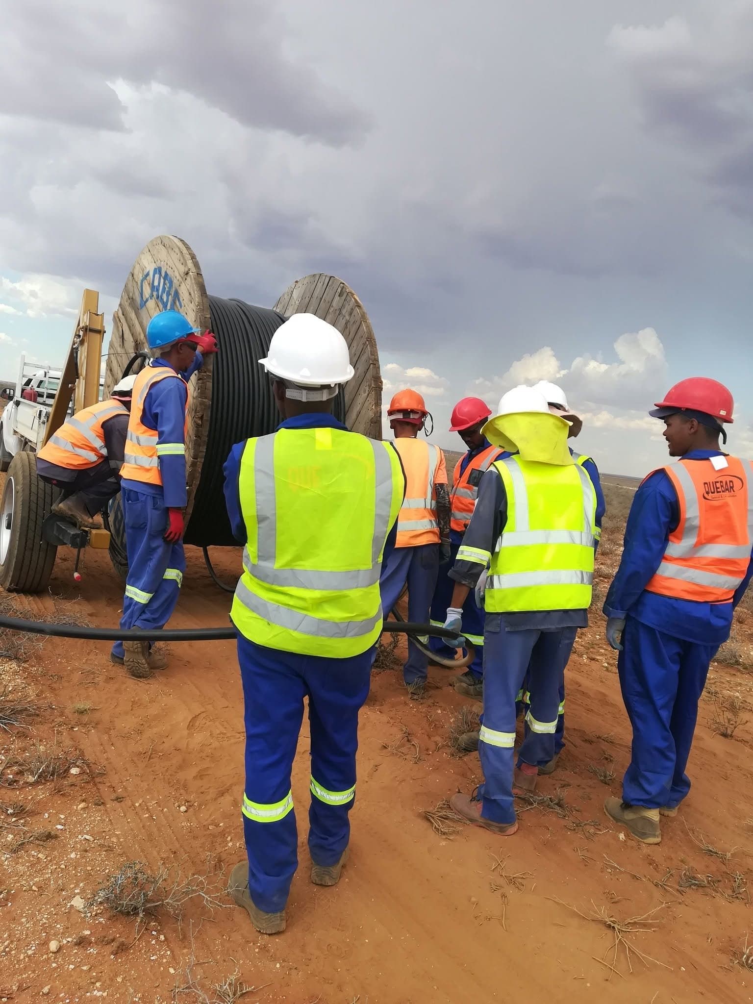 Quebar crew in hi-viz vests, hard hats and blue overalls pulling cable off a drum on a wind farm under a gathering storm
