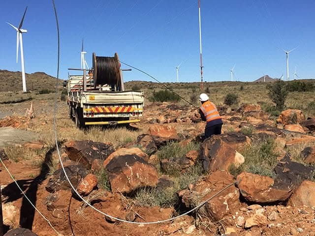Linesman in hi-viz and hard hat pulling conductor from a drum truck across rocky ground with wind turbines behind